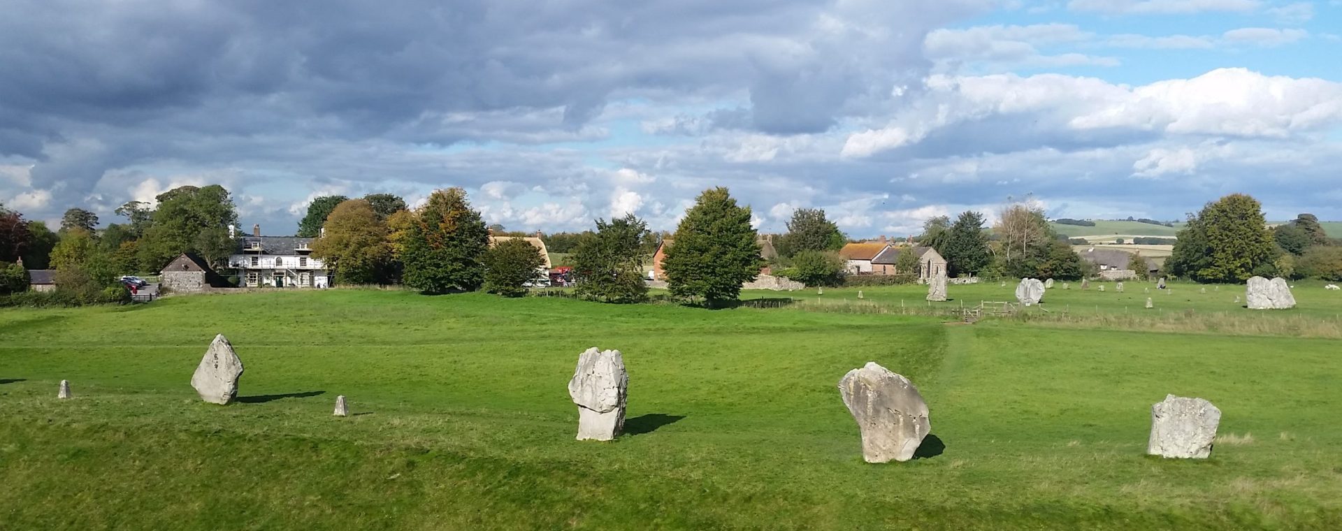 A view across Avebury stones on a fresh spring day. Ancient stones rise from the fresh green grass, trees and picturesque village buildings in the distance.